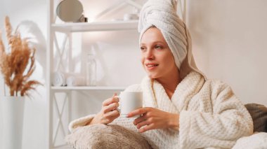 Spa sabahı. Çay gevşemesi. Tazelik ilhamı. Bornozlu mutlu bir kadın banyodan kupa üstüne kupa ile dinleniyor açık ev iç mekanında fotokopi alanı ile..