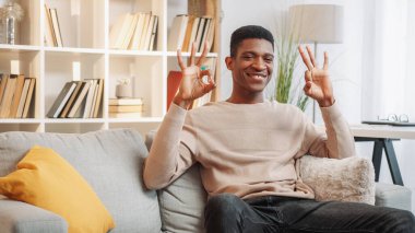 Onaylandı. Tamam jest. Memnuniyet verici genç adam evdeki boş alandaki oturma odasındaki rahat koltukta işaret dili gösteriyor..