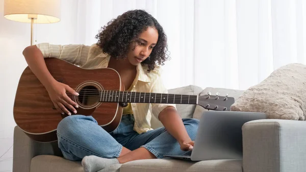 Online guitar lesson. Music education. Home leisure. Creative woman learning string instrument at laptop practicing chords on couch light room.