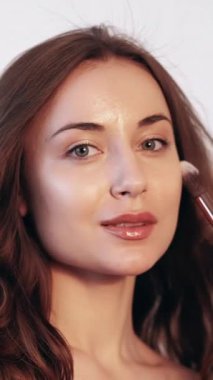 Dikey video. Günlük makyaj. Temeli uygula. Yüz hatları. Profesyonel bir yüz ifadesi. Kusursuz cildinde pudra fırçası olan genç bir kadın..