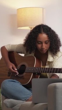 Dikey video. Gitar alıştırması. Çevrimiçi müzik dersi. Karantina eğitimi. Mutlu kadın gitarist kanepe odasında dizüstü bilgisayar dersi alıyor..