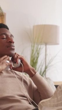 Dikey video. Şarkı sevinci. Müzik aleti. Ses eğlencesi. Kulaklıkla en sevilen müzik listesini dinleyen mutlu adam evdeki oturma odasında ellerini kaldır..