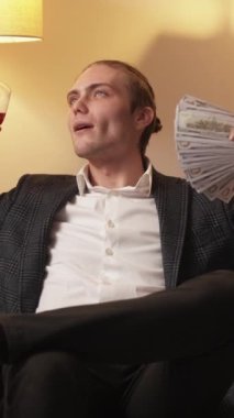 Dikey video. Zenginler lüks bir hayat. Zenginlik başarısı. Memnun milyoner iş adamı dolar para ile rahatlığın tadını çıkarıyor. Kanepede kırmızı şarap içiyor..