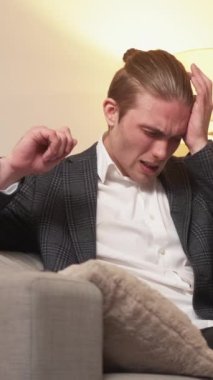 Dikey video. İş yorgunluğu. Stres gerginliği. Sağlıksız fazla çalışma. Evdeki kanepede dinlenirken baş ağrısı çeken yorgun bir adam..