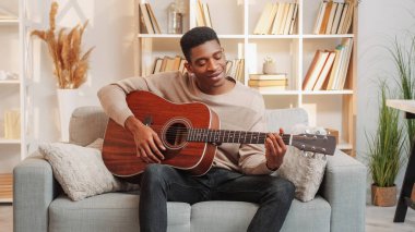 Gitar alıştırması. Müzisyen eğlencesi. Yaratıcı hobi. Yetenekli, neşeli bir adam evdeki oturma odasındaki kanepede telli çalgılarla şarkı söylüyor..