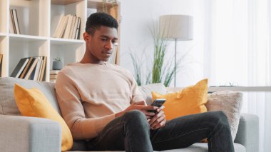 Haberci sohbeti. Gezici eğlence. Çevrimiçi boş zaman. Rahatlamış genç adam akıllı telefondan sosyal medyada mesaj atıyor evdeki oturma odasındaki kanepede internete bakıyor..