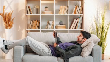 Cihaz dinlendirici. İnternet eğlencesi. Hafta sonu evde. Dijital tablette sörf yapan mutlu bir adam oturma odasında rahat bir kanepenin keyfini çıkarıyor..