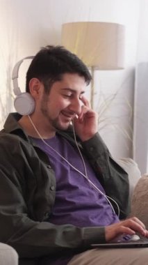 Dikey video. Müzik dinliyor. Cihaz eğlencesi. Ev eğlencesi. Mutlu adam kulaklıkla en sevdiği şarkıyı dinliyor. Oturma odasındaki kanepede dizüstü bilgisayarında şarkı listesi yapıyor..