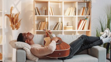 Gitar dinlendirici. Müzikal eğlence. Ev eğlencesi. Huzurlu rüya gibi adam akustik telli enstrüman ile şarkı akorları çalıyor. Oturma odasındaki rahat kanepede hafta sonunun tadını çıkarıyor..