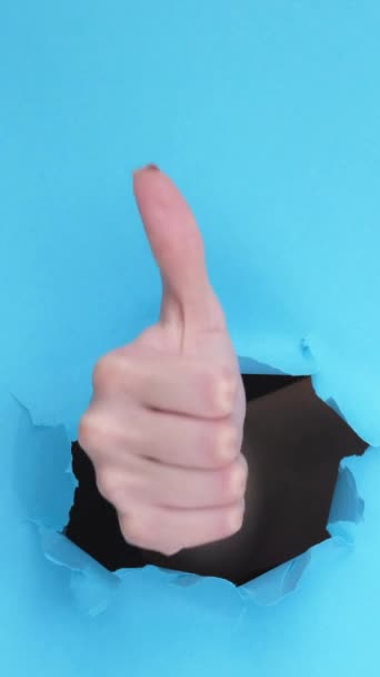 垂直录像 像手一样完美的选择 在有空隙的蓝色背景上 通过突破纸撕破墙壁的孔来接受 的拇指向上举的姿态 — 图库视频影像