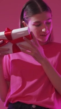 Dikey video. Doğum günü hediyesi. Tatil hediyesi. Şenlik sürprizi. Pasifik Pembe Neon ışıklı arka planda paketlenmiş mutlu kadın.