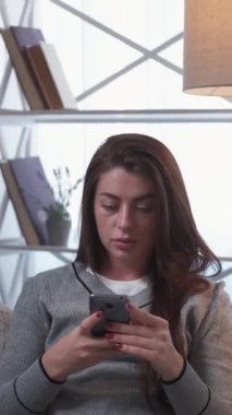 Dikey video. Mesafe çalışması. Konuşan kadın. Mobil teknoloji. Konsantre bayan kanepede oturuyor. Kitap açtı. Işıklandırma odasının içinde akıllı telefon yazdı..