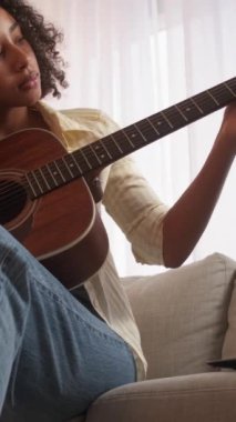 Dikey video. Gitar hobisi. Müzik alıştırması. Hafta sonu tatili. Odaklanmış bir kadın akustik telle akorlar çalıyor. Oturma odasında koltukta oturuyor..