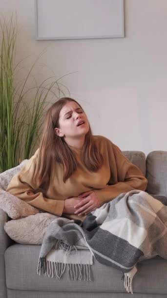垂直录像 肚子痛 Belly抽筋了身体不健康的妇女感到剧痛 双手紧握着肚子坐在沙发上 躺在光线充足的房间里 — 图库视频影像