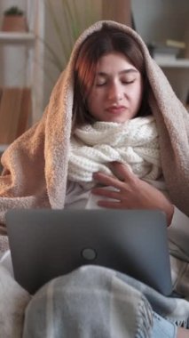 Dikey video. Hastalıklı iş. Grip bağımlısı. Bitkin, titreyen bir kadın ekoseli bir bilgisayarla çevrimiçi iş bilgisayarına sarılmış. Oturma odasındaki iç kanepede kendini rahatsız hissediyor..
