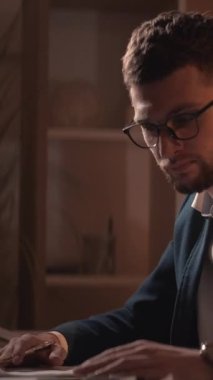 Dikey video. İş başarısız oldu. Endişeli adam. Kayıp belgeler. Meşgul adam kendini çaresiz hissediyor. Kağıt çalışıyor. Karanlık odada masa başında oturuyor..
