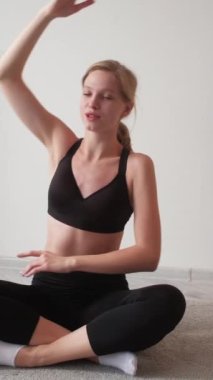 Dikey video. Ev pilatesi. Yoga egzersizi. Kapalı spor salonu eğitimi. Rahatlamış atletik kadın sırt kaslarını esnetiyor yerde bağdaş kurarak oturuyor..