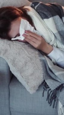 Dikey video. Ateşim çıktı. Grip hapşırması. Sağlıksız kız kanepede peçete gribi semptomları göstererek yatıyor..