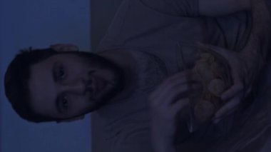 Dikey video. Film gecesi. Ev eğlencesi. Keyifli adam, komik komedi dizilerini izlerken eğleniyor. Mavi ışıklı karanlık odada cipsleri yerken gülüyor..