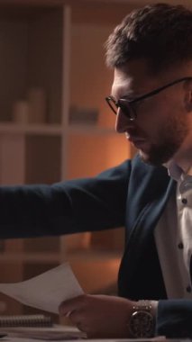 Dikey video. Bilgileri. Endişeli adam. İş başarısız oldu. Gerekli belgeleri bulmak için kendini çaresiz hisseden meşgul bir adam karanlık odanın içinde masa başında oturuyor..