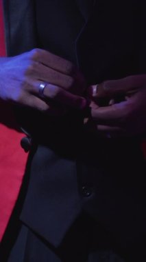 Dikey video. Centilmen tarzı. Şık bir moda. Lüks parti görünüşü. Neon ışığı tanınmayan adam koyu mor üzerine kırmızı şık smokin ilikliyor..