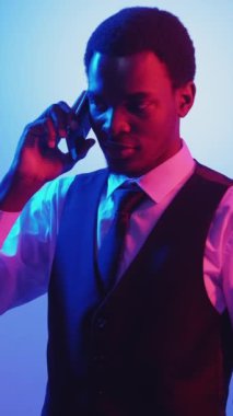 Dikey video. İş görüşmesi. Neon ışığı insanları. Mobil iletişim. Kendine güvenen güler yüzlü adam telefonda pembe ışıkta mavi üzerine konuşuyor..