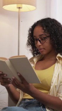 Dikey video. Ev ödevi. Tembel hafta sonu. Odaklanmış düşünceli kadın öğrenci kitap hikayesinin keyfini çıkarıyor, aydınlık odanın içini rahatlatıyor..