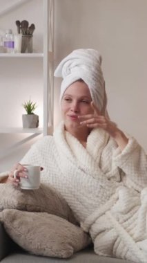 Dikey video. Sabah kahvesi. Haftasonu rahatlama. Spa eğlencesi. Bornozlu mutlu bir kadın sıcak içkinin keyfini çıkarıyor. Açık ev iç kısımlarında banyo üstüne kanepede hayal kuruyor..