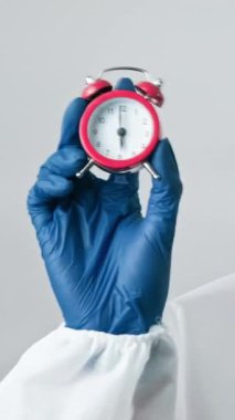 Dikey video. Tam zamanında teşhis kondu. Virüs uzmanı. Gri arka planda çalar saati olan tıbbi koruyucu ekipmanlı kadın doktor..