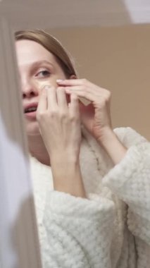 Dikey video. Göz bantları. Cilt bakımı ürünü. Cilt tazeleme. Altın kolajen hidrojenin aynadaki yansımasına yüz pedleri takan kendinden emin sarışın kadın..