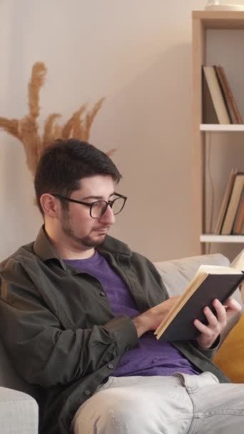 垂直ビデオ 家の読書 余暇を予約する 週末はリラックス 快適なソファの上で居心地の良いリビングルームで文学を楽しむ眼鏡の平和的な男 — ストック動画