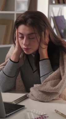 Dikey video. Migren ağrısı. Hasta kadın. Mesafe çalışması. Yorgun bir kadın kafasında dayanılmaz bir ağrı hissediyor. Masasında dizüstü bilgisayarı ve iç mekânında oturuyor..