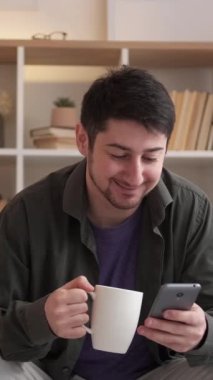 Dikey video. Gezici sohbet. Hücre eğlencesi. Dijital iletişim. Neşeli bir adam akıllı telefondan mesaj atıyor evde kahve içiyor, dinleniyor..