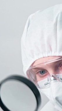 Dikey video. Viroloji araştırması. Tıbbi muayene. Loupe 'un gri arka planda izole edilmiş koruyucu giysi ekipmanları konusunda uzman kadın doktor..