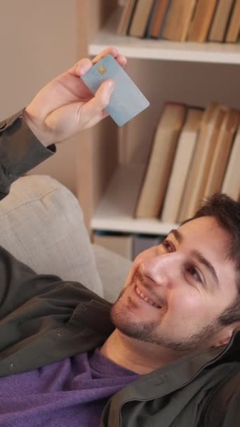 垂直录像 信用卡服务 银行客户 塑料借记 快乐的男人在沙发上放松一下 满足于在网上购物 还能在家里用现金还钱 — 图库视频影像
