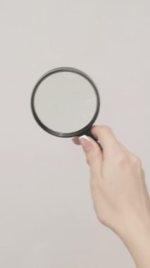 Dikey video. Yakınlaştırıcı cam. Mercek keşfediliyor. Gri arkaplanda izole edilmiş büyüteçle boş alanı inceleyen kadın eli.