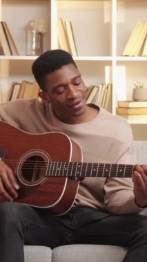 Dikey video. Gitar alıştırması. Müzisyen eğlencesi. Yaratıcı hobi. Yetenekli, neşeli bir adam evdeki oturma odasındaki kanepede telli çalgılarla şarkı söylüyor..