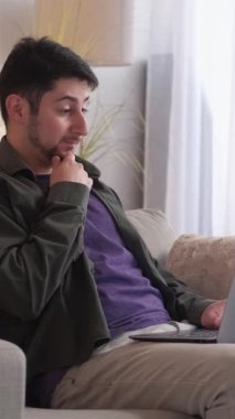 Dikey video. Düşünceli bir çalışan. İş problemi çözümü. Uzak geliştirici. Kafası karışmış adam evdeki kanepede dizüstü bilgisayarda proje araştırması yapıyor..