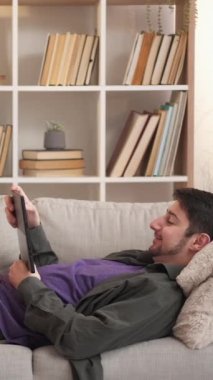 Dikey video. Cihaz dinlendirici. İnternet eğlencesi. Hafta sonu evde. Dijital tablette sörf yapan mutlu bir adam oturma odasında rahat bir kanepenin keyfini çıkarıyor..
