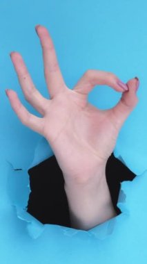Dikey video. Tamam jest. Mükemmel seçim. Mavi yırtık duvardaki kağıt deliğinin içinde onaylanmış parmak dairesi işareti gösteren bir kadın el..