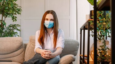 Ev izolasyonu. Karantina hijyeni. Koruyucu tıbbi maske takan güzel bir kadın pandemik süreçte yalnız başına kanepede oturuyor..