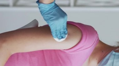 Dikey video. Covid-19 aşısı. Kas içi enjeksiyon. Koronavirüs salgını. Eldivenli hemşire, maske takmış hastayı pamuklu bezle siliyor ve bantlıyor..