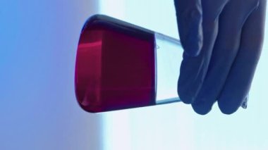 Dikey video. Klinik test. Kimya laboratuvarı. İlaç denemesi. Mavi eldivenli teknisyen eli kırmızı çözeltili cam matara reaksiyonu kontrol ediyor.
