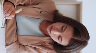 Dikey video. İnternet terapisi. Online oturum. Psikolog işi. Takım elbiseli düşünceli bir kadın beyaz iç mekânda notlar yazarken başını sallıyor..