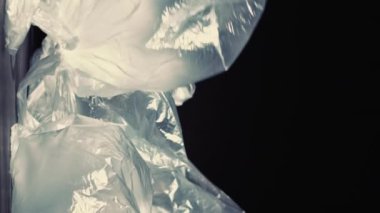 Dikey video. Atık toplama. Plastik geri dönüşüm. Çevre kirliliği. Şişmiş polietilen paketler siyah arka planda izole edilmiş çöp yığınından yükseliyor..