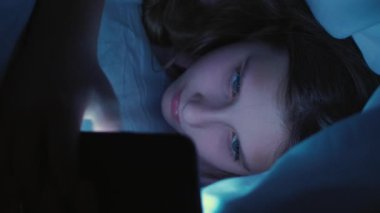 Dikey video. Ekran zamanı. Çocuk uykusuzluğu. Geç saate kadar. İnternet bağımlılığı. Uykusuz, yorgun kız akıllı telefondan mesajlaşıyor evde battaniyenin altında saklanıyor..