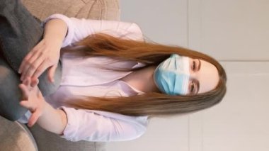 Dikey video. İnternet günlüğü. Salgın konuşmacı. Koruyucu tıbbi maskeli bir kadın kanepeye oturup karantina tavsiyesi veriyor..