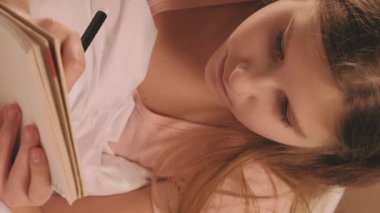 Dikey video. Çocuk günlüğü. Yaratıcı hobi. Çocuk hayal kuruyor. Eskiz defterinden ilham alan kız, sabahları evde rahat beyaz bir battaniyenin içinde dinleniyor..
