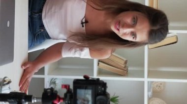 Dikey video. Vlog kaydı. İçerik oluştur. Kadın bir uzman. Genç duygusal dışavurumcu kadın tripodla kameraya konuşuyor ve evde iç mekanda el kol hareketi yapıyor..