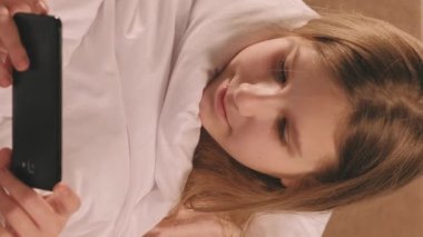 Dikey video. Dijital çocukluk. Çevrimiçi eğlence. Mutlu gülümseyen mutlu kız, sabahları battaniyenin altında komedi programı seyrediyor..